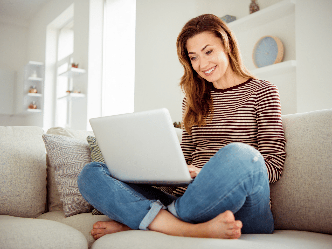 Close up photo of an attractive lady holding a notebook and Skypes relatives friends wearing jeans and striped pullover while sitting on cosy divan house living indoors. Communications Concept.