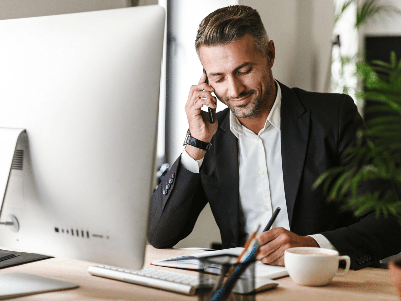 Image of handsome businessman talking on cell phone while working on computer in office. Contact Concept.