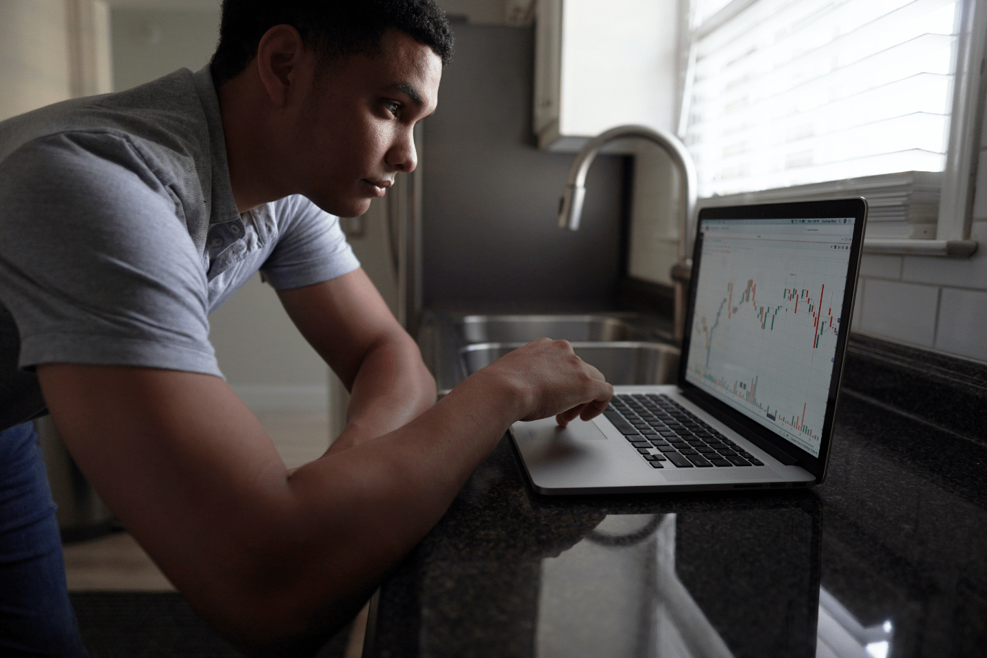 Handsome young African American man leaning over laptop placed upon a black granite countertop in a modern kitchen. He is looking at a stock chart. Portfolio monitoring/ Save More Money concept.