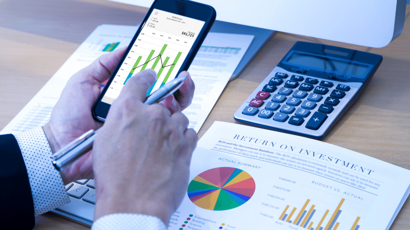 Investment Performance and Risk Analysis: Businessman touching smart phone application in front of modern desktop computer analyzing return on investment. Risk Versus Return Concept.