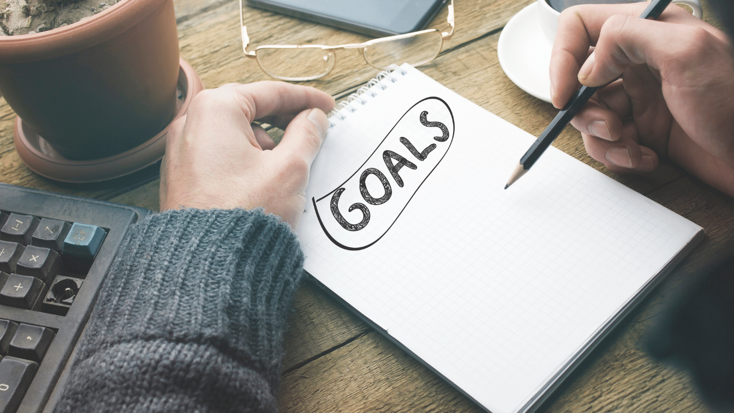 Financial Goals Concept: Person at a wooden desk writing on a notebook that says 