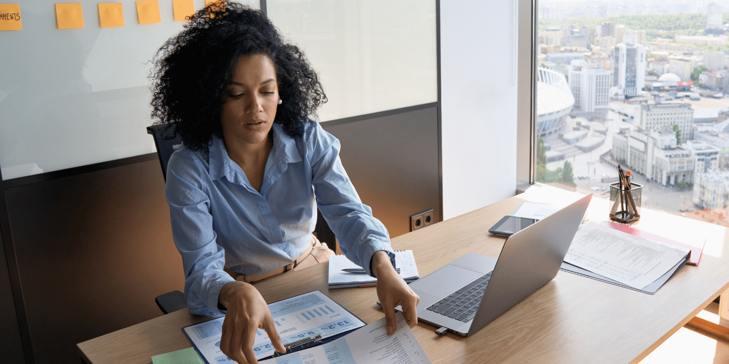 African American Businesswoman CEO analyst sitting at desk working with papers with laptop near panoramic window. Financial Goals Concept.