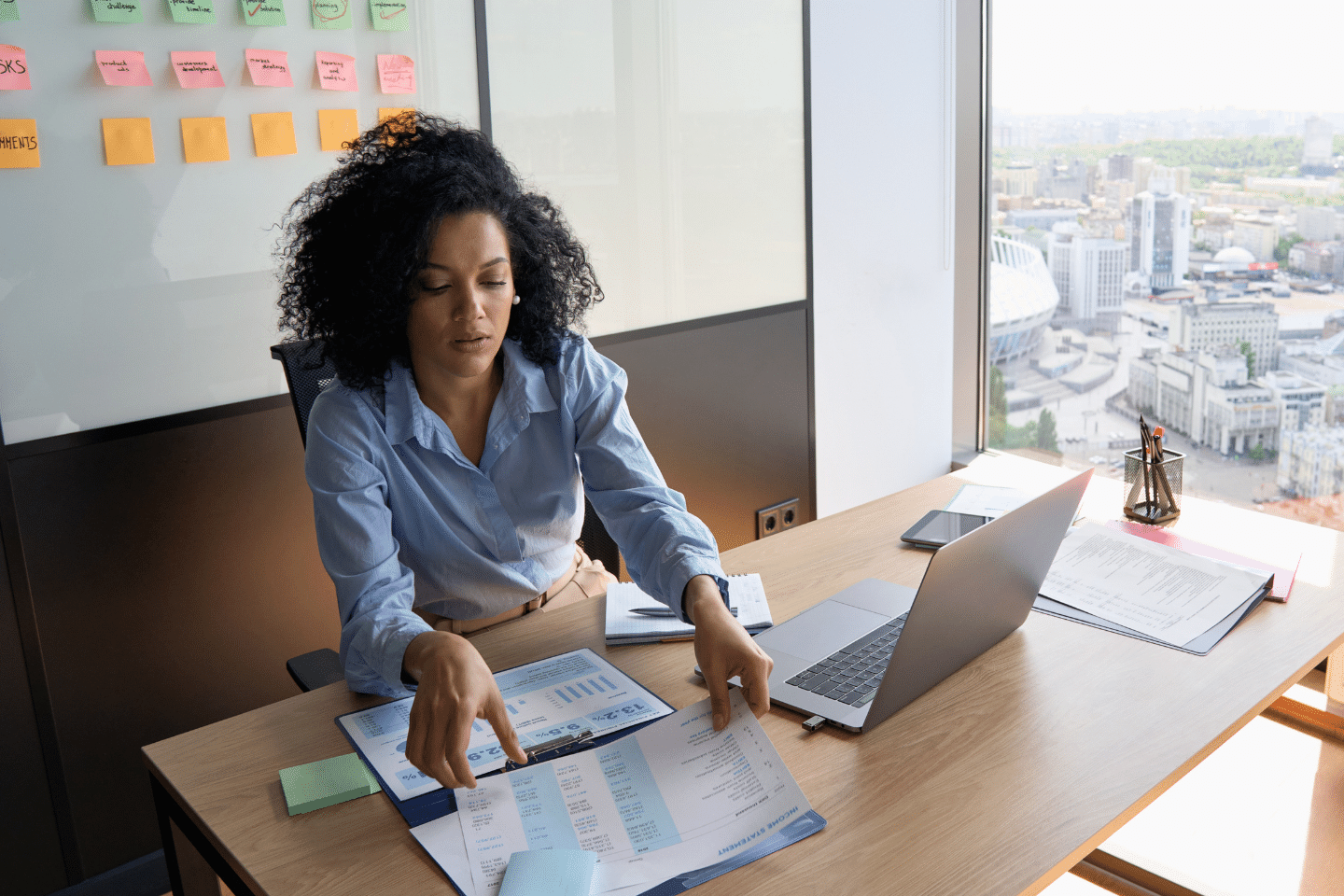 African American Businesswoman CEO analyst sitting at desk working with papers with laptop near panoramic window. Financial Goals Concept.