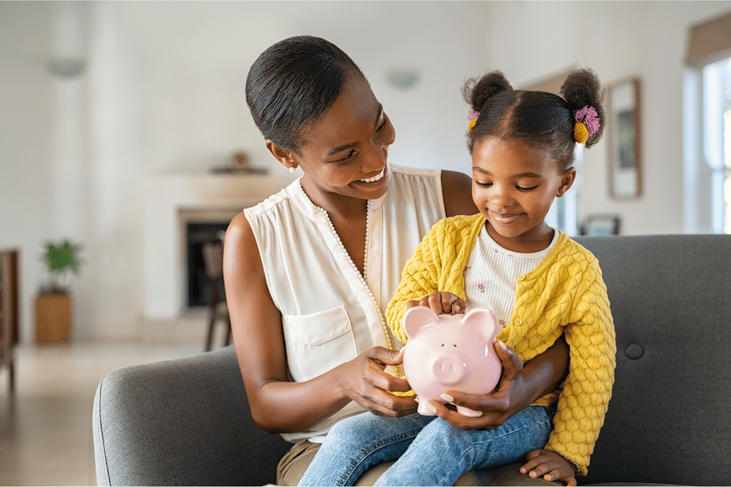 Black Woman with Daughter Holding Piggy Bank: Smiling mature African American woman helping daughter sitting on lap putting money in a piggy bank. Cute little Black girl. Saving Concept.